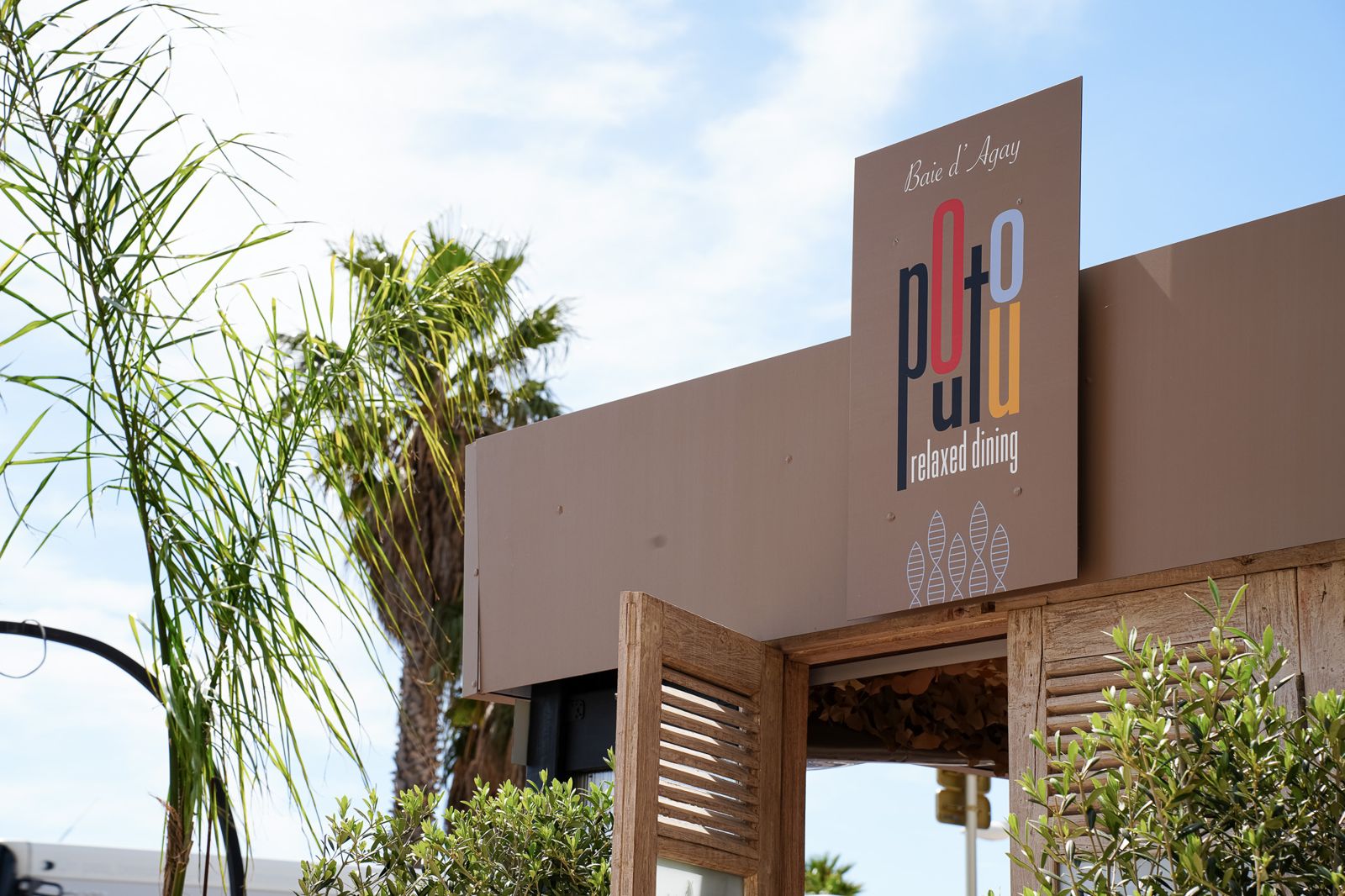 L'entrée du restaurant, devant un grand panneau « Baie d'Agay, poutou, relaxed dining » sous un ciel bleu et un palmier
