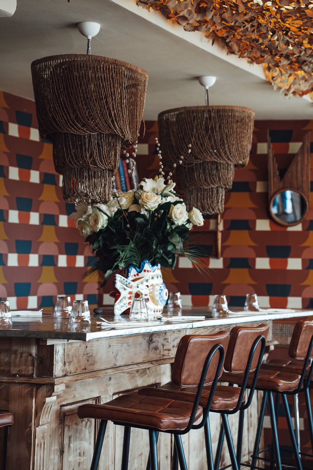 Le bar du restaurant devant un mur de carreaux géométriques terracotta, bleu et moutarde, suspensions en fibres et bouquet de fleurs