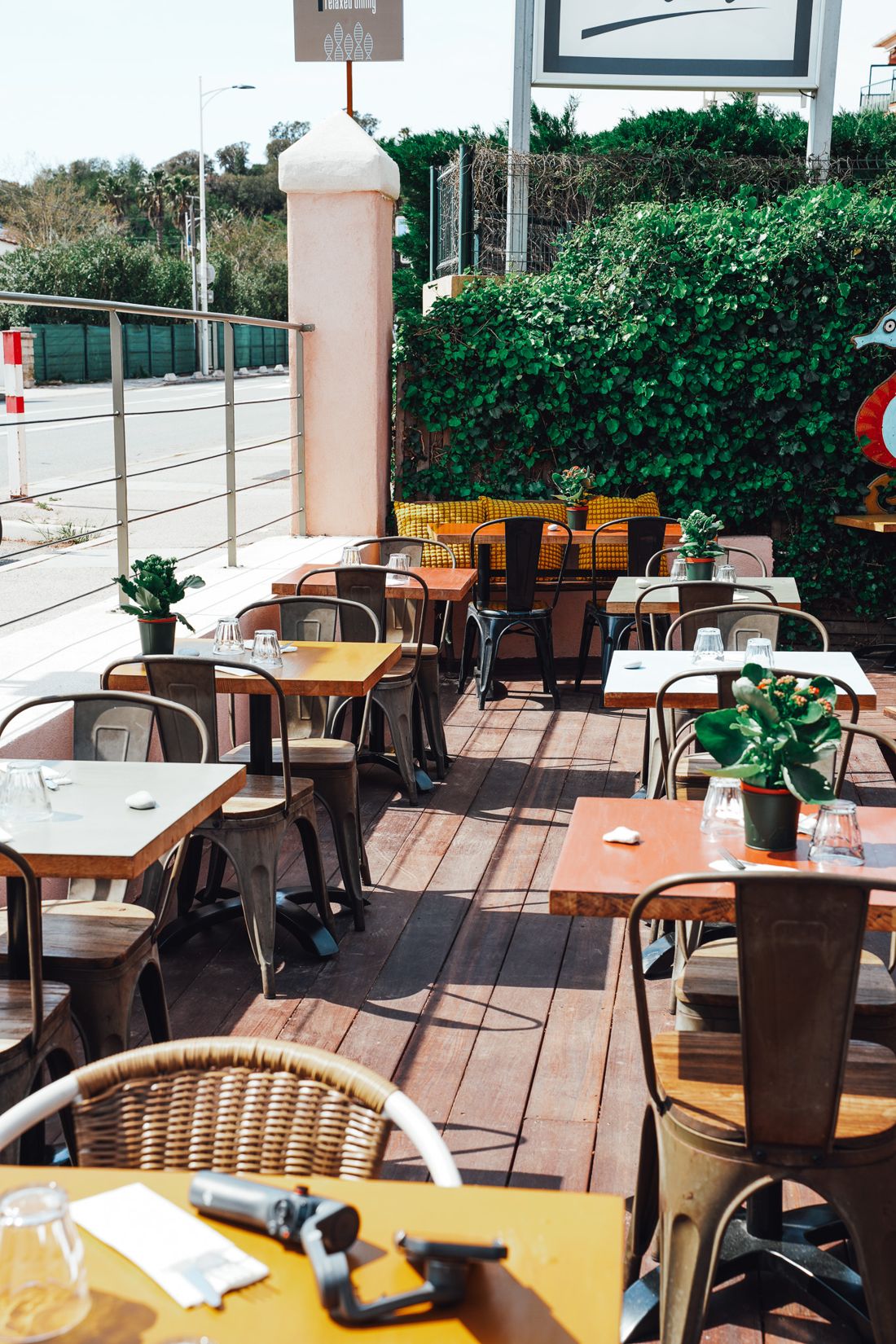 La terrasse ensoleillée : tables en bois, chaises en métal et en rotin sur un plancher, haies vertes